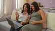© Krakenimages.com - Mother and pregnant daughter smiling and waving while making a video call in the living room of their home, capturing a warm family moment indoors with love and togetherness.
