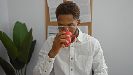 Young man drinking coffee in an office setting, sporting glasses, white shirt, and surrounded by indoor plants and bulletin board with papers.