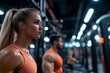 © MaMaKe - A young woman in an orange tank top looks determined as she works out in a gym, with other people working out in the background.
