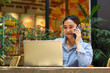 © M Alfan Setyawan - happy asian woman enjoy working on laptop and talking on mobile phone while sitting in outdoor cafe with green plant interior