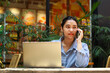 © M Alfan Setyawan - happy asian woman enjoy working on laptop and talking on mobile phone while sitting in outdoor cafe with green plant interior