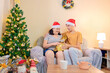 © Marcio - A small family exchanging Christmas gifts at home, wearing Santa hats by a decorated tree. A warm moment between a mother and her adult son, highlighting holiday traditions and joy