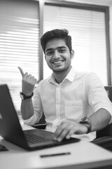 Man is sitting at a desk with a laptop in front of him. He is smiling and pointing to the laptop screen