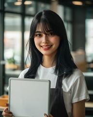 young Asian woman in a modern office setting wearing a white t-shirt and dark apron, holding a medium size whiteboard with blank screen, 