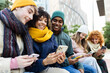 © Xavier Lorenzo - Diverse group of young friends using mobile phones sitting on a city bench. Multiracial Gen Z student people in warm clothes browsing social media and connecting online. Youth and technology concept