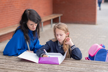 smart girl solving math problems on her own while her mother sits nearby at wooden table on children's playground and watches,education concept,going back to school, girl in beautiful blue uniform