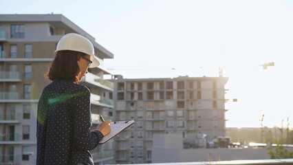 Woman architect wearing a hard hat is taking notes with a tablet computer while inspecting a building construction site at sunset