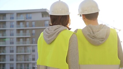 Man and woman engineers wearing safety hard hats and vests holding blueprint and discussing something on construction site at sunrise, back view. Architecture and engineering concept