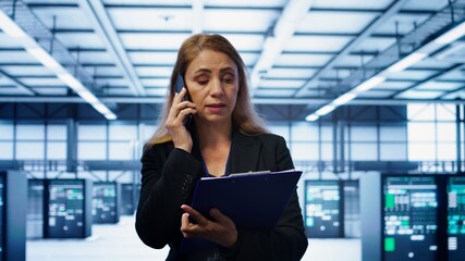 IT specialist looking over data center maintenance checklist, talking on phone in server farm. Employee surrounded by server racks talking during telephone call, camera B