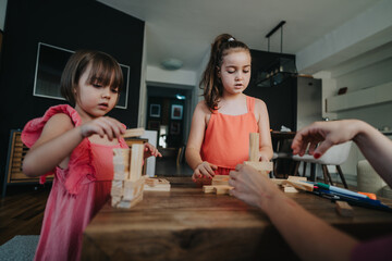 Two young girls engaged in creative play with wooden blocks at a home table. Focused and imaginative learning through building and teamwork in a cozy family setting.