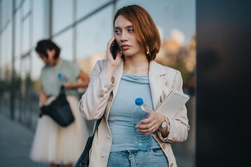 A young businesswoman in casual attire engages in a phone call outside a modern glass building, portraying urban life and professionalism. She holds a water bottle and notebook, depicting multitasking