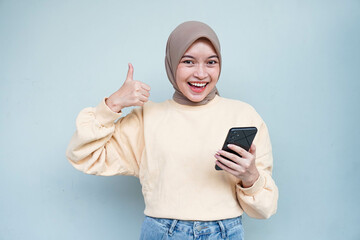 Smiling young Asian Muslim woman holding mobile phone and making thumb up gesture isolated over white background.