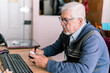 © Centric  - Senior man in glasses writing in a notebook next to his computer at a home office