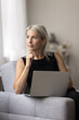 © fizkes - Pensive older age female working on distance at home office sit on couch look aside distracted from laptop screen. Aged remote worker create new idea for email marketing ponder on job. Vertical image