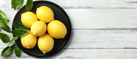 Fototapete - Fresh ripe juicy lemons on a black plate with green leaves on a white wooden table from a top view copyspace for your text