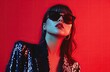 © thiquynh - Portrait of a fashionable woman in neon light, wearing stylish sunglasses and a dark blue sequin suit. The fashion model poses isolated on a red background with copy space