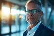 © Odin AI - A middle-aged man wearing glasses stands with confidence in a modern office setting, his face illuminated by the warm glow of the sun through large windows.