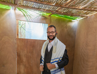 Smiling jew man wearing a traditional Jewish prayer shawl (tallit) and formal attire, standing inside a wooden sukkah with a roof made of leaves. Religious or cultural moment with the natural light