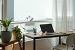 © DragonImages - Urban home office setup featuring a minimalist desk, laptop, and potted plants next to large window allowing soft daylight to fill room, creating calming atmosphere
