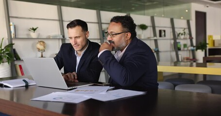Two multiethnic business professionals collaboratively analyze data on laptop, sitting at desk in coworking office reviewing charts and graphs to develop strategic solutions for their joint project