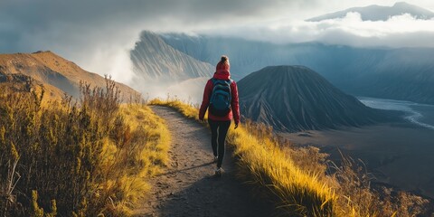 A person is walking on a path in the mountains. The person is wearing a red jacket and a backpack. The mountains are covered in trees and the sky is cloudy