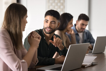 Handsome young Middle Eastern company intern talking to coworker woman at laptop, discussing corporate professional education, training task. Two colleagues, partners working at computer together