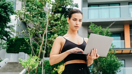 A young woman types on her laptop near a modern building, focused on her tasks. An ideal spot for creativity and inspiration in an urban setting.