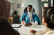 © pressmaster - Muslim female doctor in clinic office giving medical advice to patient, holding medication bottle. Stethoscope around neck, medical documents on desk, indoor environment