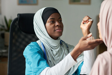 Muslim female doctor examining patient with stethoscope in a modern medical office. She is smiling warmly at the patient and wearing professional medical attire