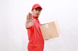 © ArvStd - Asian delivery courier in red t-shirt and cap carrying a box package while making stop gesture showing palm of hand. isolated over white background
