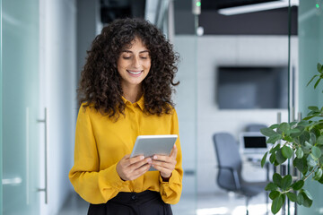 Latin American businesswoman in office using tablet, representing professionalism. Smiling in yellow blouse, standing in hallway with digital device, symbolizing technology in workplace.