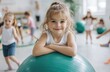 © thiquynh - Cute little girl sitting on a green fitness ball in the children's gym, smiling and looking at the camera with her arms folded, surrounded by other kids doing various sports activities