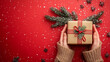 © Martinesku - Female's hands holding Christmas gift box decorated with evergreen branch on red background with snow. Christmas and New Year. Banner concept.