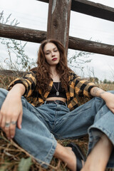 Wall Mural - Fashion beautiful autumn fresh girl in fashionable clothes with shirt, vintage blue jeans sits in the grass near a wooden fence countryside in nature