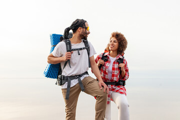 young african american couple tourists with hiking backpacks and equipment traveling and walking near sea, woman and man standing near lake and looking at horizon