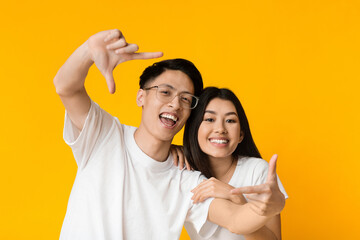 Asian man and woman laughing and showing frame sign with their hands on yellow studio background