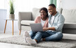 © Prostock-studio - Happy black lovers sitting on floor on carpet by couch with laptop, having fun together at home, copy space. Cheerful african american couple looking for new furniture online, websurfing