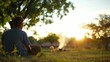 © MaMaKe - A man sits in a field, gazing towards the sunset.  He is surrounded by trees and grass, and there is a small fire in the distance.