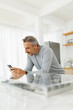 © eggeeggjiew - Middle aged man using a smartphone with a focused expression, dressed in a gray sweater in a bright, modern kitchen interior.