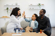 © sofiko14 - African female doctor gives high five to young boy while sitting with father at home. Warm interaction promotes trust and comfort in home healthcare setting.