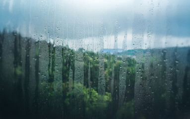 Rain streaks down a window, blurring the view of a lush green landscape.