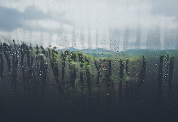 Poster - Rain water droplets cover a window pane, blurring the view of a lush, green landscape with mountains in the background.