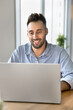 © fizkes - Positive handsome freelance professional man in casual working on online project at laptop, typing, smiling, browsing Internet, social media, sitting at workplace table in home office. Vertical shot