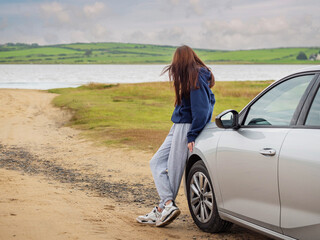 A woman is sitting on the side of the road next to a silver car. She is wearing a blue hoodie and gray sweatpants. The scene is quiet and peaceful. Model looking at calm nature scene. Travel to nature