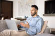 © fizkes - Handsome young Hispanic man talking on work conference chat at laptop, resting on home sofa, speaking on video call at computer, holding computer on lap, using pc gadget for job communication