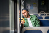 Thoughtful man concentrating on mobile phone rides on electric train reading e-book, scrolling social media, listens to music on headphones to pass time, leaning to window. Public transport trip alone