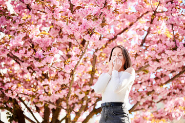 Woman allergic suffering from seasonal allergy at spring in blossoming garden at springtime. Woman sneezing and blowing nose using nasal handkerchief in front of blooming tree. Spring allergy concept.