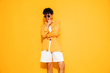 Young attractive man with short curly hair hairstyle. Serious handsome male in casual stylish shirt and shorts clothes posing in studio, isolated on yellow. Cheerful and happy model. In his thoughts