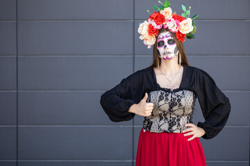 woman sugar skull makeup design floral crown gives thumbs up gray wall. wearing black top and red skirt. Day of the Dead holiday traditionally celebrated on November 1 and 2. October 31 or November 6