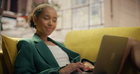 Poster - Young Black Woman Reclines on Yellow Couch, Using Laptop in Bright Room. She Types and Smiles. Her Green Blazer Contrasts With the Couch. Sunlight Streams Through Large Windows. Slow Motion